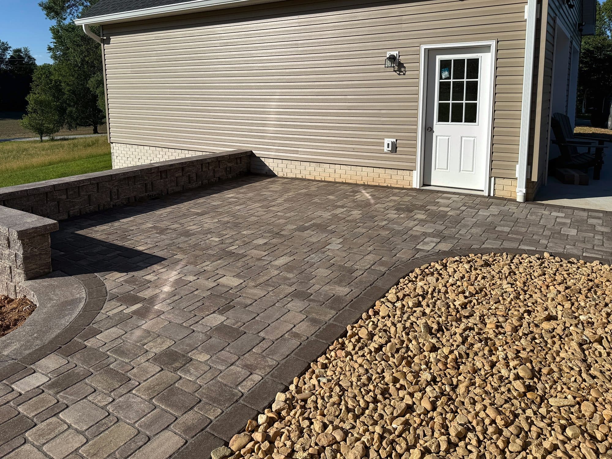 Tumbled paver patio with a segmental seat wall and a curved river rock bed beside a garage entry.