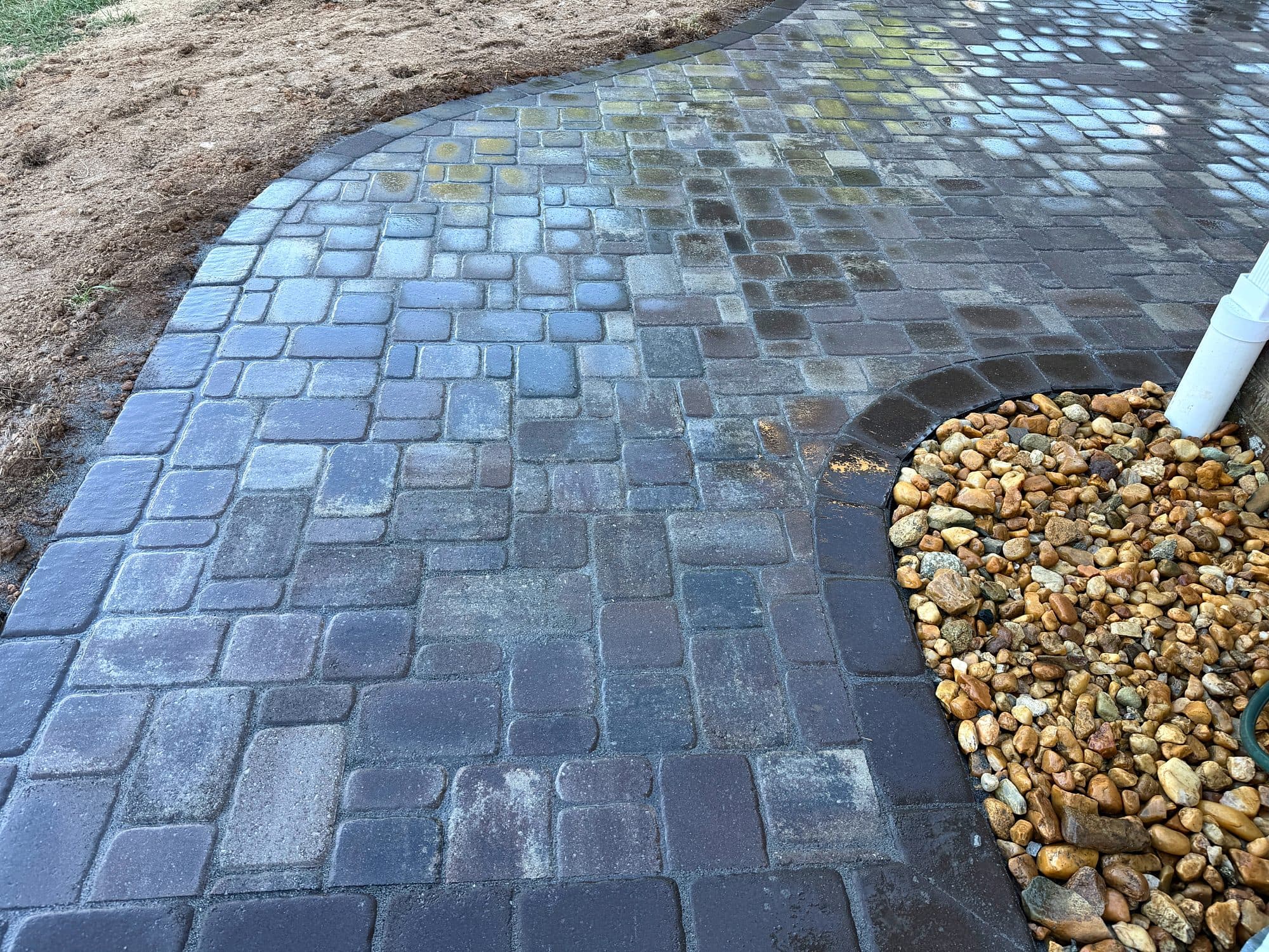Close view of a curved tumbled-paver edge with a soldier-course border around a river rock downspout bed.